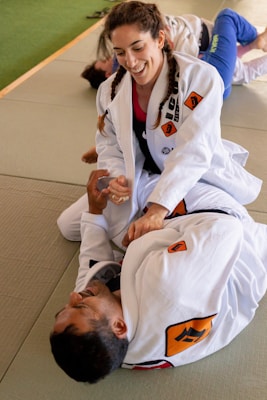 Two people are practicing Brazilian Jiu-Jitsu on a mat, wearing white gis with various patches. One person, smiling with braided hair, is in a top position, appearing to demonstrate a technique on the other person lying on their back.