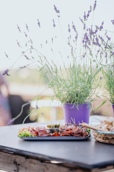 A table is set with a tray of charcuterie including meats, pickles, and garnishes, surrounded by potted lavender plants in purple pots. There is also a basket of sliced bread. The setting appears casual and outdoors, suggesting a relaxed dining experience.
