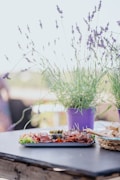 A table is set with a tray of charcuterie including meats, pickles, and garnishes, surrounded by potted lavender plants in purple pots. There is also a basket of sliced bread. The setting appears casual and outdoors, suggesting a relaxed dining experience.