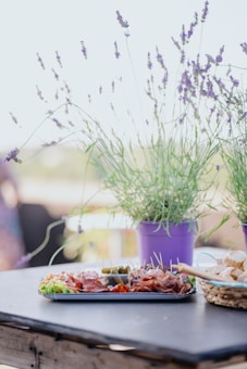 A table is set with a tray of charcuterie including meats, pickles, and garnishes, surrounded by potted lavender plants in purple pots. There is also a basket of sliced bread. The setting appears casual and outdoors, suggesting a relaxed dining experience.