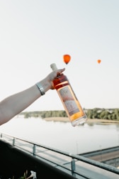 A hand holding a bottle of rosé wine is prominently displayed against a serene background of a river and distant trees. Three hot air balloons, two in the air and one closer to the water, are visible in the sky.