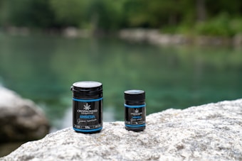 Two containers labeled 'Crystalweed Amnesia' are placed on a large rock in an outdoor setting. A tranquil, blurred background of greenery and water suggests a natural environment, possibly near a river or lake.