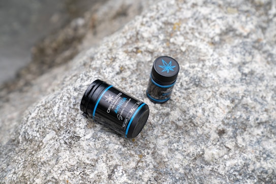 Two black cylindrical containers with blue accents and a cannabis leaf design lie on a textured stone surface. One container is slightly larger than the other, and both display labels suggesting cannabis-related products.