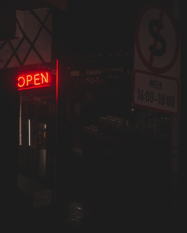 A dimly lit scene featuring a bright red neon 'OPEN' sign on the left side. To the right, a sign with time restrictions is visible, indicating a 'No Parking' symbol with specified hours from 16:00 to 18:00. The background remains dark, with hints of shelves or industrial elements barely visible.