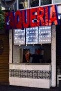 A street food stand with a large sign reading 'TAQUERIA' in bold red and blue letters. Below the sign is a menu listing various types of tacos and prices. A person wearing a red hat is seen inside, working with a vertical rotisserie, preparing meat. The counter features decorative tiles with a floral pattern.