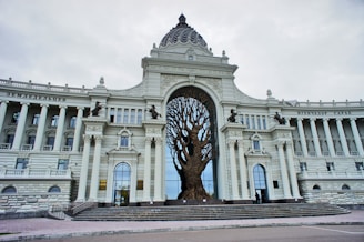 A grand architectural building featuring classical elements with columns and arches. At the center, a large sculpture of a tree with expansive branches is integrated into the structure. The facade is adorned with intricate details and statues of seated figures.