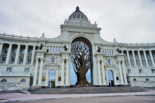 A grand architectural building featuring classical elements with columns and arches. At the center, a large sculpture of a tree with expansive branches is integrated into the structure. The facade is adorned with intricate details and statues of seated figures.