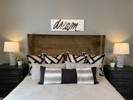 A neatly arranged bedroom features a plush bed with a soft beige blanket and a variety of decorative pillows in black, white, and gray patterns. Above the bed, a minimalist sign with the word 'dream' is mounted on the wall. Two bedside tables flank the bed, each holding a modern table lamp with white shades and a small potted plant. The room is anchored by a large, padded headboard in a rich brown color.