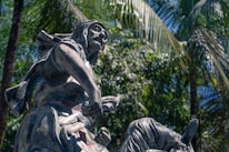 A dynamic sculpture of a human figure in motion, set against a lush background of tropical palm trees. The figure appears to be in a dramatic pose, possibly with a weapon in hand. The sculpture's detailed clothing and expressive face convey a sense of action and history.