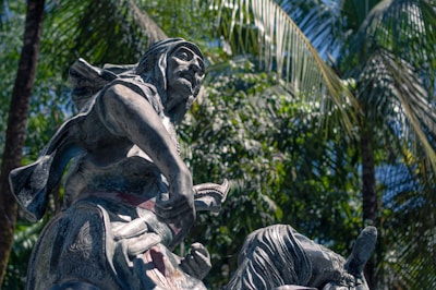 A dynamic sculpture of a human figure in motion, set against a lush background of tropical palm trees. The figure appears to be in a dramatic pose, possibly with a weapon in hand. The sculpture's detailed clothing and expressive face convey a sense of action and history.