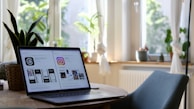 A laptop is placed on a round wooden table in a brightly lit room. The laptop screen displays comparisons between two social media platforms. The room has large windows with white curtains letting in plenty of natural light. There are green plants on the windowsill and a wicker plant pot on the table. A blue chair is positioned at the table.