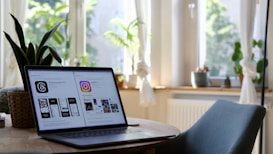 A laptop is placed on a round wooden table in a brightly lit room. The laptop screen displays comparisons between two social media platforms. The room has large windows with white curtains letting in plenty of natural light. There are green plants on the windowsill and a wicker plant pot on the table. A blue chair is positioned at the table.