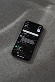 A modern smartphone displaying a social media app interface is lying on a textured gray surface. The screen shows a post by a user followed by comments and interaction icons like like, comment, and share. The ambient lighting casts a soft shadow around the phone.