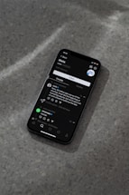 A modern smartphone displaying a social media app interface is lying on a textured gray surface. The screen shows a post by a user followed by comments and interaction icons like like, comment, and share. The ambient lighting casts a soft shadow around the phone.