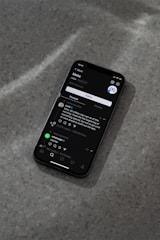 A modern smartphone displaying a social media app interface is lying on a textured gray surface. The screen shows a post by a user followed by comments and interaction icons like like, comment, and share. The ambient lighting casts a soft shadow around the phone.