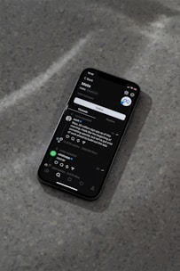 A modern smartphone displaying a social media app interface is lying on a textured gray surface. The screen shows a post by a user followed by comments and interaction icons like like, comment, and share. The ambient lighting casts a soft shadow around the phone.