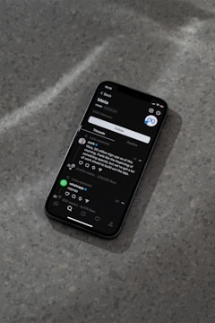 A modern smartphone displaying a social media app interface is lying on a textured gray surface. The screen shows a post by a user followed by comments and interaction icons like like, comment, and share. The ambient lighting casts a soft shadow around the phone.