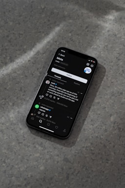 A modern smartphone displaying a social media app interface is lying on a textured gray surface. The screen shows a post by a user followed by comments and interaction icons like like, comment, and share. The ambient lighting casts a soft shadow around the phone.