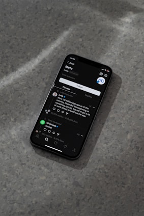 A modern smartphone displaying a social media app interface is lying on a textured gray surface. The screen shows a post by a user followed by comments and interaction icons like like, comment, and share. The ambient lighting casts a soft shadow around the phone.