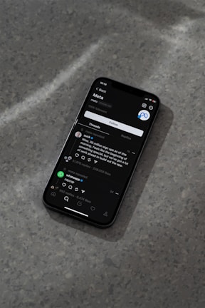 A modern smartphone displaying a social media app interface is lying on a textured gray surface. The screen shows a post by a user followed by comments and interaction icons like like, comment, and share. The ambient lighting casts a soft shadow around the phone.