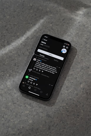 A modern smartphone displaying a social media app interface is lying on a textured gray surface. The screen shows a post by a user followed by comments and interaction icons like like, comment, and share. The ambient lighting casts a soft shadow around the phone.