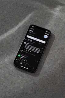 A modern smartphone displaying a social media app interface is lying on a textured gray surface. The screen shows a post by a user followed by comments and interaction icons like like, comment, and share. The ambient lighting casts a soft shadow around the phone.