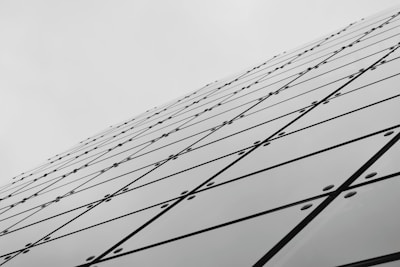 A series of reflective glass panels arranged in a geometric pattern, creating a dynamic diagonal line that extends toward the sky. The panels are secured with bolts, and each panel features intersecting lines forming a grid-like appearance, contributing to a modern architectural aesthetic.