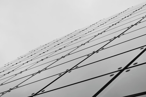 A series of reflective glass panels arranged in a geometric pattern, creating a dynamic diagonal line that extends toward the sky. The panels are secured with bolts, and each panel features intersecting lines forming a grid-like appearance, contributing to a modern architectural aesthetic.