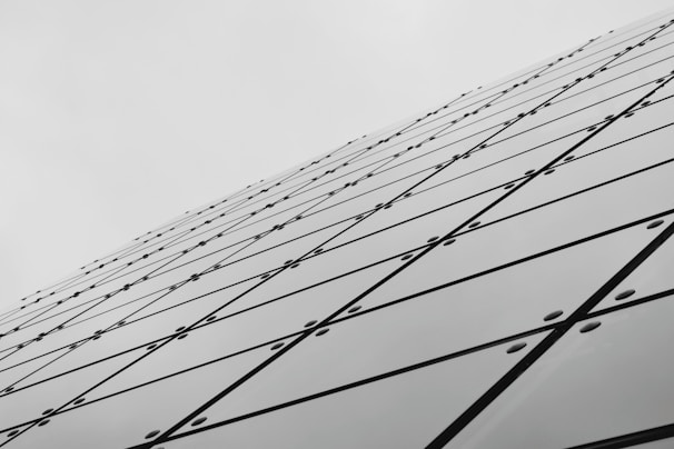 A series of reflective glass panels arranged in a geometric pattern, creating a dynamic diagonal line that extends toward the sky. The panels are secured with bolts, and each panel features intersecting lines forming a grid-like appearance, contributing to a modern architectural aesthetic.