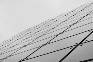 A series of reflective glass panels arranged in a geometric pattern, creating a dynamic diagonal line that extends toward the sky. The panels are secured with bolts, and each panel features intersecting lines forming a grid-like appearance, contributing to a modern architectural aesthetic.