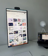 The image features a modern desk setup with a vertically oriented large monitor displaying a web design portfolio or gallery. The screen shows multiple web design thumbnails. There is a keyboard and mouse on the white desk surface, and a laptop is situated to the right, slightly open and displaying different content. A round white lamp is positioned in the background, adding a minimalist touch to the overall workspace.