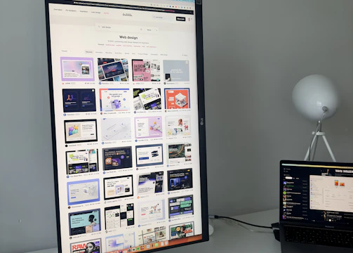 The image features a modern desk setup with a vertically oriented large monitor displaying a web design portfolio or gallery. The screen shows multiple web design thumbnails. There is a keyboard and mouse on the white desk surface, and a laptop is situated to the right, slightly open and displaying different content. A round white lamp is positioned in the background, adding a minimalist touch to the overall workspace.