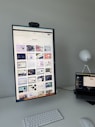 The image features a modern desk setup with a vertically oriented large monitor displaying a web design portfolio or gallery. The screen shows multiple web design thumbnails. There is a keyboard and mouse on the white desk surface, and a laptop is situated to the right, slightly open and displaying different content. A round white lamp is positioned in the background, adding a minimalist touch to the overall workspace.