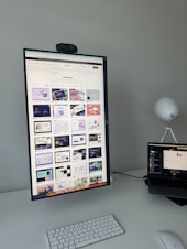 The image features a modern desk setup with a vertically oriented large monitor displaying a web design portfolio or gallery. The screen shows multiple web design thumbnails. There is a keyboard and mouse on the white desk surface, and a laptop is situated to the right, slightly open and displaying different content. A round white lamp is positioned in the background, adding a minimalist touch to the overall workspace.