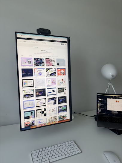 The image features a modern desk setup with a vertically oriented large monitor displaying a web design portfolio or gallery. The screen shows multiple web design thumbnails. There is a keyboard and mouse on the white desk surface, and a laptop is situated to the right, slightly open and displaying different content. A round white lamp is positioned in the background, adding a minimalist touch to the overall workspace.