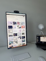 The image features a modern desk setup with a vertically oriented large monitor displaying a web design portfolio or gallery. The screen shows multiple web design thumbnails. There is a keyboard and mouse on the white desk surface, and a laptop is situated to the right, slightly open and displaying different content. A round white lamp is positioned in the background, adding a minimalist touch to the overall workspace.