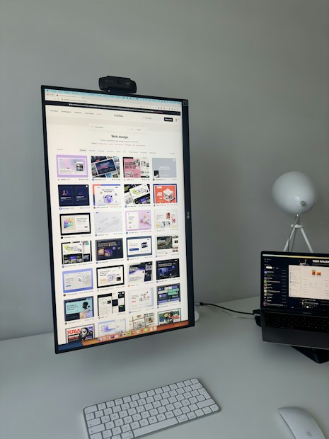 The image features a modern desk setup with a vertically oriented large monitor displaying a web design portfolio or gallery. The screen shows multiple web design thumbnails. There is a keyboard and mouse on the white desk surface, and a laptop is situated to the right, slightly open and displaying different content. A round white lamp is positioned in the background, adding a minimalist touch to the overall workspace.