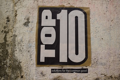 A textured wall features a large poster with the words 'TOP 10' in bold black and white lettering. Below the main text, a smaller phrase reads 'solutions for the common good'.
