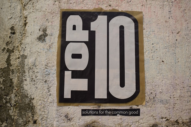 A textured wall features a large poster with the words 'TOP 10' in bold black and white lettering. Below the main text, a smaller phrase reads 'solutions for the common good'.