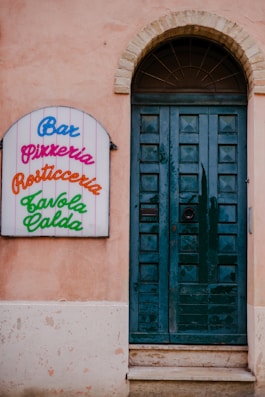 A rustic building facade features a dark teal wooden door with multiple panels, framed by a light stone arch at the top. To the left of the door, there is a colorful sign with the words 'Bar', 'Pizzeria', 'Rosticceria', and 'Tavola Calda' written in various bright colors. The exterior wall is painted a faded peach color with visible weathering.