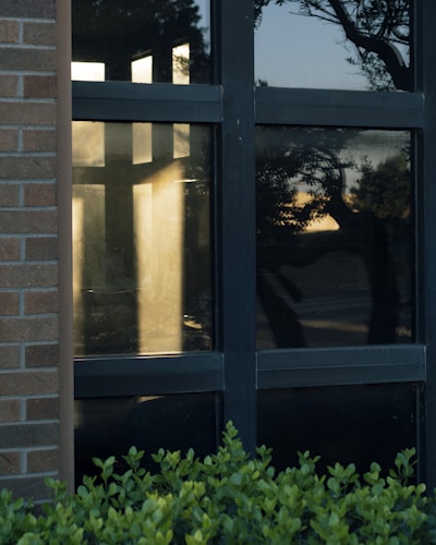Reflection of sunlight on a large cross-shaped window pane surrounded by dark framing. The window is part of a building with a brick exterior. There are green shrubs in front of the window and a silhouetted view of trees and a sunset sky is visible through the glass.