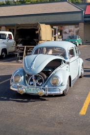 A vintage light blue Volkswagen Beetle is parked in an outdoor lot with its hood open, revealing the spare tire and part of the engine. The vehicle is clean and well-maintained, with distinctive chrome hubcaps and yellow-tinted headlight covers. In the background, a few other classic vehicles and a building with a beige facade and a sloped roof are visible.
