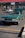 A classic green convertible car with its hood open is parked in a shopping center parking lot. The front of the car is prominently displayed, showing the vintage design with circular headlights. There is another car parked in the background near some storefronts with a red and green sign above.