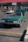A classic green convertible car with its hood open is parked in a shopping center parking lot. The front of the car is prominently displayed, showing the vintage design with circular headlights. There is another car parked in the background near some storefronts with a red and green sign above.