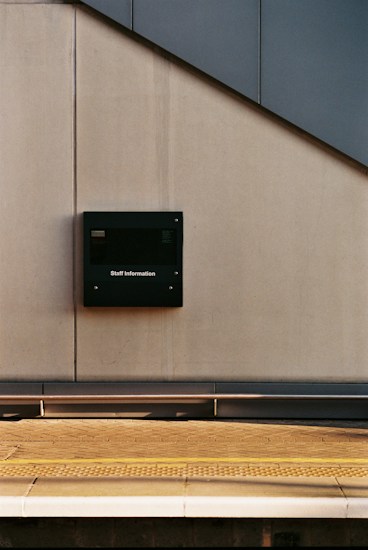 A minimalist scene featuring a train station platform. A light gray wall with a dark information board marked 'Staff Information' is visible. The platform edge is highlighted with a textured yellow safety strip. The angled sunlight creates long shadows and a calm atmosphere.