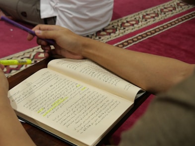 An open book with Arabic text is held by a person who is holding a purple pen and a yellow highlighter. The background features a red carpet with a decorative pattern, and part of another person sitting nearby is visible.