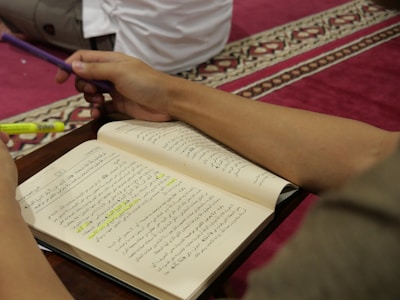 An open book with Arabic text is held by a person who is holding a purple pen and a yellow highlighter. The background features a red carpet with a decorative pattern, and part of another person sitting nearby is visible.
