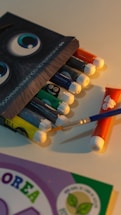 A set of watercolor paint tubes arranged next to each other. Each tube is a different color, and a blue paintbrush rests on the surface next to them. The image also features the corner of an open paintbox with a character illustration on it and a partially visible booklet or packaging in the bottom corner.