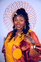 A woman stands confidently, adorned in a vibrant traditional dress featuring a combination of yellow and brown fabric. Her hair is styled in long curls, accessorized with a decorative clip. She is smiling warmly, wearing large hoop earrings and a necklace. A detailed henna design is visible on her hand, complemented by gold bangles on her wrist. Behind her, a decorative circular pattern adds to the composition.