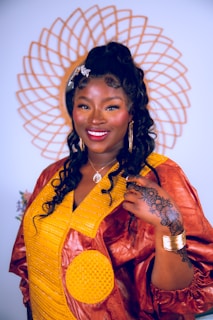 A woman stands confidently, adorned in a vibrant traditional dress featuring a combination of yellow and brown fabric. Her hair is styled in long curls, accessorized with a decorative clip. She is smiling warmly, wearing large hoop earrings and a necklace. A detailed henna design is visible on her hand, complemented by gold bangles on her wrist. Behind her, a decorative circular pattern adds to the composition.